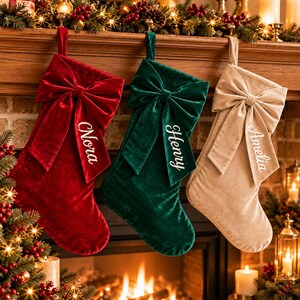 May include: Three velvet Christmas stockings in red, dark green, and beige, each featuring a large decorative bow at the top. The stockings are personalized with white embroidered names: Nora on the red, Henry on the green, and Amelia on the beige.