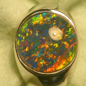 Op de afbeelding: Een zilveren ring met een ronde, veelkleurige opaal-edelsteen die in het midden is gezet. De opaal heeft een donkerblauwe basis met flitsen van groen, rood en geel.