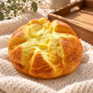 Könnte beinhalten: Ein rundes, goldbraun gebackenes Brötchen mit einer kreuzförmigen Vertiefung auf der Oberseite. Die Kruste wirkt strukturiert und leicht knusprig, während das Innere einen helleren, blassgelben Kern mit einer weichen, porösen Struktur zeigt.