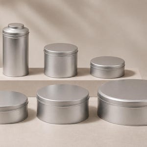 May include: A collection of six silver-coloured metal canisters and round tins of varying heights and diameters. The set includes one tall cylindrical canister with a lid and five flat, circular containers with removable lids arranged in a group.