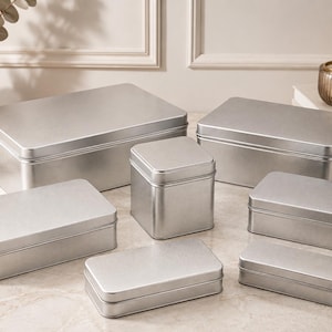 Op de afbeelding: Een set van zeven rechthoekige en vierkante zilverkleurige metalen blikken met afneembare deksels. De set bevat verschillende formaten, variërend van een grote rechthoekige doos tot kleinere, smalle rechthoekige blikken en één vierkant blik.
