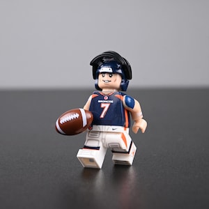 Puede incluir: Una minifigura de jugador de fútbol americano de los Denver Broncos con una camiseta azul marino con el número 7, pantalones blancos con detalles naranjas y un casco negro. La figura sostiene un balón de fútbol marrón con cordones blancos.