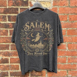 Peut inclure: Un t-shirt gris anthracite à manches courtes avec une impression graphique portant le texte "SALEM 1692" et "They Missed One". Le motif représente une sorcière sur un balai, un croissant de lune, des étoiles, une ville et des fleurs.