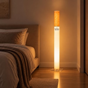 May include: A tall, cylindrical floor lamp designed to resemble a cigarette. The top section features an orange, speckled filter pattern, while the lower white body displays the black text Marlboro MEDIUM. It stands on a clear square base.