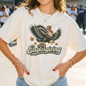 Könnte beinhalten: Ein cremefarbenes Kurzarm-T-Shirt mit einer Grafik eines Truthahns mit ausgebreiteten Flügeln, der über einem Football steht und von herbstlichen Blättern umgeben ist. Darunter steht das Wort Eaglesgiving in dunkelgrüner Schreibschrift.