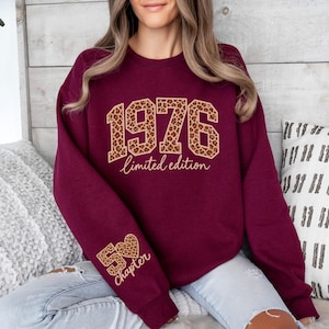 May include: A maroon crewneck sweatshirt featuring the text 1976 in a leopard print pattern with the words limited edition underneath. The right sleeve displays the text 50 chapter with a heart icon, also in a leopard print pattern.