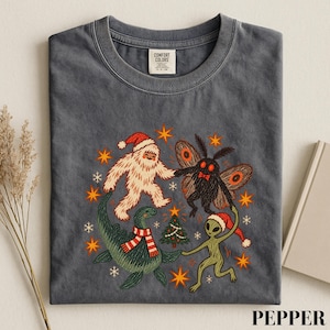 May include: A charcoal gray t-shirt featuring a graphic of a white yeti in a Santa hat, a mothman, a green sea monster in a striped scarf, and a green alien in a Santa hat dancing around a small Christmas tree with orange stars and snowflakes.