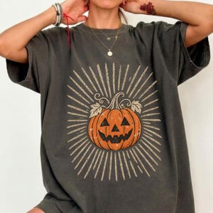 May include: A charcoal gray short-sleeve t-shirt featuring a central graphic of an orange jack-o'-lantern pumpkin. The pumpkin is surrounded by a radiating sunburst pattern composed of thin, light-colored lines and small leopard print spots.