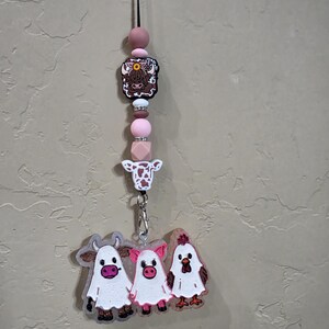 May include: A hanging car charm featuring a black cord with pink, white, and brown beads, a highland cow bead, and a cow head bead. A silver lobster clasp connects to a clear acrylic charm displaying a cow, a pig, and a chicken, each wearing a white ghost costume.