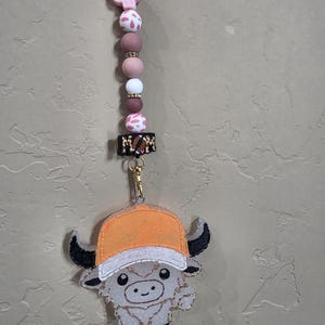 May include: A hanging car charm featuring a beaded strand with a pink cactus, white and pink cow print beads, solid mauve beads, and gold spacers. A black rectangular bead reads MOM with a football icon. A glittery white highland cow wears an orange cap.