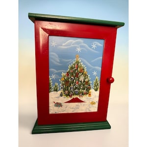 May include: A rectangular wooden wall cabinet with a red frame and green top and bottom trim. The center panel features a painted scene of a decorated Christmas tree in a snowy landscape with white snowflakes against a blue sky.