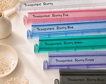 Puede incluir: Una colección de seis soportes de exhibición rectangulares hechos de material translúcido con detalles de purpurina. Los soportes están etiquetados como Transparent Starry, Transparent Starry Pink, Transparent Starry Blue, Transparent Starry Green, Transparent Starry Purple y Transparent Starry Black.