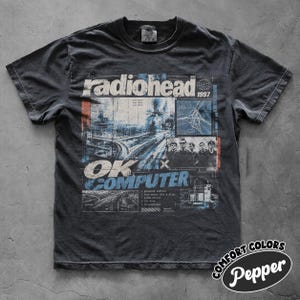 May include: A charcoal gray short-sleeve t-shirt featuring a distressed graphic print. The design includes the text radiohead, 1997, and OK COMPUTER, alongside a train track illustration, a band photograph, and technical grid elements.