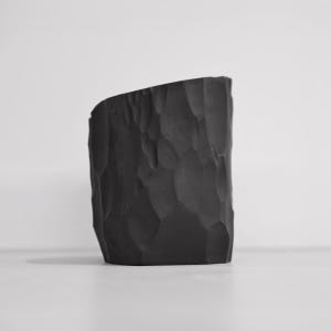 May include: A matte black cylindrical vessel featuring a hand-carved, faceted exterior surface with irregular indentations. The top edge of the container is cut at a slight angle, creating a non-uniform rim profile that contrasts with the dark finish.