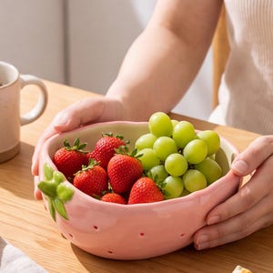 Pode incluir: Uma tigela de cerâmica rosa claro em forma de morango com detalhes de folhas verdes na lateral, cheia de morangos vermelhos e uvas verdes frescas. Uma pequena estatueta de cerâmica de um gato segurando uma fatia de melancia está próxima.