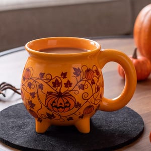Puede incluir: Una taza de cerámica de color naranja brillante con forma de caldero, tres patas pequeñas y un asa curva. El exterior presenta un diseño de líneas de color marrón oscuro con calabazas y hojas de parra que envuelven el cuerpo redondeado.