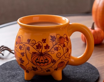 Könnte beinhalten: Ein leuchtend orangefarbener Keramikbecher in Kesselform mit drei kleinen Füßen und einem geschwungenen Henkel. Die Außenseite ist mit einem dunkelbraunen Linienmuster aus Kürbissen und geschwungenen Weinblättern rund um den Körper verziert.