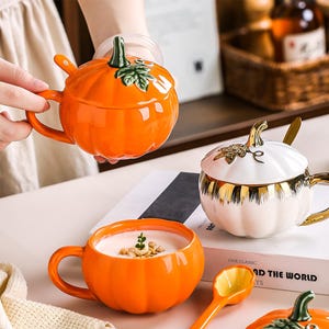 May include: A collection of ceramic pumpkin-shaped mugs with lids and matching spoons. One mug is bright orange, another is white with gold accents, and a third orange mug is filled with milk and cereal. Each features a stem-shaped handle.