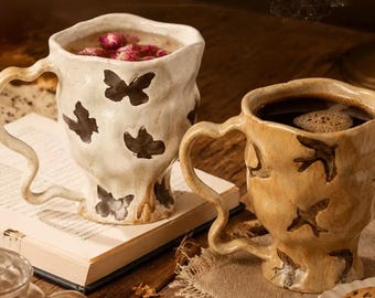 May include: Two ceramic mugs with irregular, wavy surfaces and curved handles. The left mug is off-white with dark butterfly silhouettes, filled with pink rosebuds. The right mug is tan with dark bird silhouettes, filled with black coffee.