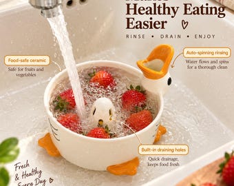 Puede incluir: Un cuenco colador de cerámica blanca con forma de pato lleno de agua y fresas frescas bajo un grifo. El cuenco tiene pies y pico de color naranja, con superposiciones de texto que dicen Makes Healthy Eating Easier, Rinse, Drain, Enjoy, Food-safe ceramic, Auto-spinning rinsing y Built-in draining holes.