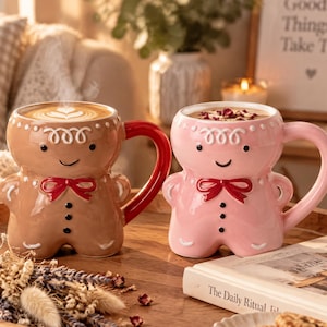 Könnte beinhalten: Zwei Keramiktassen in Form von Lebkuchenmännchen. Die linke Tasse ist braun mit einem roten Henkel, die rechte Tasse ist hellrosa mit einem passenden rosa Henkel. Beide haben weiße Glasurdetails, schwarze Augen und eine rote Fliege als Motiv.