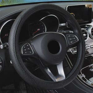 May include: A black steering wheel cover with a textured diamond pattern on the sides and smooth sections at the top and bottom. The cover is fitted onto a black steering wheel featuring integrated control buttons and silver accents in a vehicle.