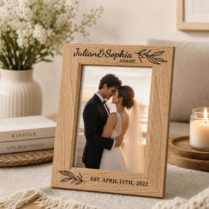 Puede incluir: Un marco de fotos rectangular de madera clara con grabado negro que dice Julian & Sophia Adams en la parte superior y EST. APRIL 15TH, 2022 en la parte inferior, con ilustraciones de hojas decorativas en los bordes del marco.
