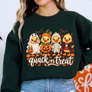 May include: A forest green long-sleeve sweatshirt featuring a graphic of four yellow ducklings in Halloween costumes, including a ghost, pumpkin, mummy, and vampire. The text below the ducks reads "quack or treat" in a stylized script font.