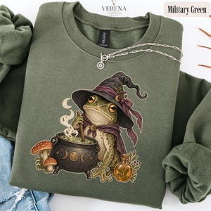 May include: A military green crewneck sweatshirt featuring a graphic of a frog wearing a purple witch hat and cape, stirring a black cauldron with moon phases. Beside the cauldron are two mushrooms and a small glass bottle filled with liquid.