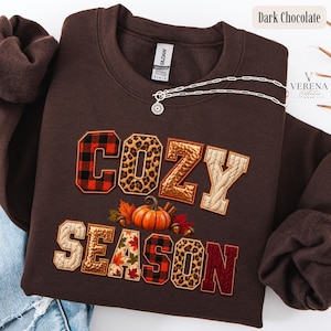 May include: A dark chocolate brown crewneck sweatshirt featuring the text COZY SEASON in varsity-style letters with various patterns including red and black buffalo plaid, leopard print, metallic gold, and cable knit, with a pumpkin and leaf graphic.