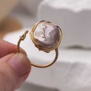Puede incluir: Un anillo con una perla barroca grande, irregular e iridiscente de color gris lavanda, engastada en una jaula de alambre fino de color dorado. La banda está hecha del mismo alambre fino, enrollado firmemente alrededor del marco.