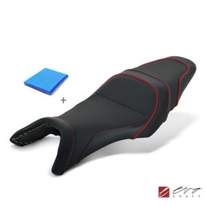 Può includere: Una sella per moto nera con profili e cuciture rosse lungo i contorni, accompagnata da un piccolo cuscinetto in gel blu rettangolare. La sella presenta un design sagomato a due sezioni con una parte anteriore stretta e una posteriore rialzata.