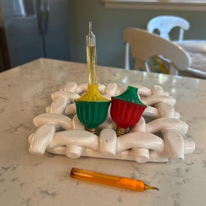 May include: Two bubble light bulbs with green and red bases, one featuring a yellow liquid-filled glass tube, resting on a white woven ceramic trivet. A separate orange liquid-filled glass tube lies horizontally in front of the trivet.