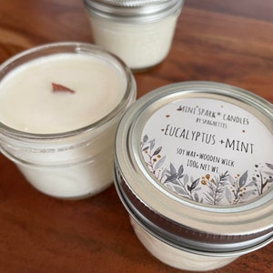 Può includere: Tre barattoli di vetro con coperchi contenenti candele di cera di soia. La candela in primo piano ha un'etichetta che recita "MINI SPARK CANDLES BY SPAGHETTIS -EUCALYPTUS +MINT SOY WAX + WOODEN WICK 100G NET WT."