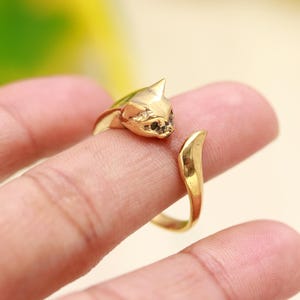 Könnte beinhalten: Ein goldfarbener offener Ring mit einem geformten Katzenkopf an einem Ende und einem spitz zulaufenden Schwanz am anderen. Die Metalloberfläche ist poliert und weist detaillierte Gesichtszüge auf, darunter spitze Ohren und dunkle Akzente.
