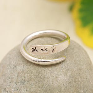May include: A silver-toned bypass ring featuring a flat, tapered end stamped with three small, dark, flower-like motifs. The band has a smooth, polished finish and a continuous, open-ended wrap design that overlaps slightly at the front.