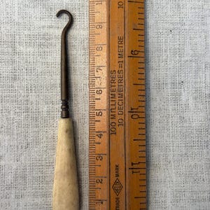 May include: A button hook with a tapered, off-white handle and a dark metal shaft ending in a curved hook. The tool is positioned next to a wooden ruler, showing the total length of the hook is approximately 11.5 cm from end to end.