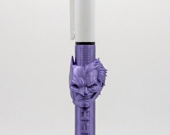 May include: A multi-color retractable ballpoint pen featuring a white top section with four colored ink selectors. The lower barrel is metallic purple with a 3D relief of a split Batman and Joker face and the word JOKER embossed vertically.
