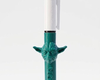 May include: A multi-color retractable ballpoint pen featuring a white upper barrel with a pocket clip and the text MADE IN FRANCE. The lower green barrel is molded with a three-dimensional head of Yoda and the embossed text YODA vertically.