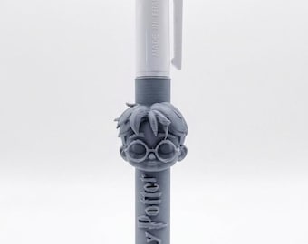 May include: A multi-color ballpoint pen with a white barrel featuring the text MADE IN FRANCE and a clip. The lower section is grey with a 3D-printed character head wearing round glasses and the embossed text Harry Potter on the grip area.
