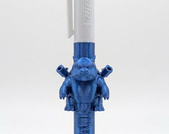 May include: A white Bic multi-color ballpoint pen featuring a metallic blue 3D-printed attachment shaped like a turtle-like creature with cannons on its back and the word TORTANK embossed vertically along the lower blue barrel section.