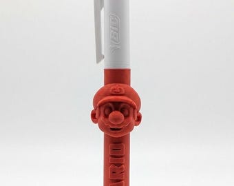 May include: A multi-color ballpoint pen featuring a white top section with the BIC logo and a red 3D-printed attachment. The attachment displays a sculpted Mario character head and the word MARIO embossed vertically along the red barrel.