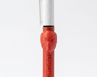 May include: A multi-color retractable ballpoint pen featuring a white upper barrel with the BIC logo and a clip. The lower barrel is red, embossed with the word SPIDERMAN, and includes a molded red plastic Spider-Man mask topper on the grip.