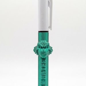 Peut inclure: Un stylo à bille rétractable multicolore avec un corps blanc portant l'inscription MADE IN FRANCE. Le stylo présente un embout vert métallique imprimé en 3D en forme de visage et le texte en relief KRAMP HATER sur la partie inférieure.