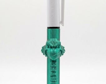 May include: A multi-color retractable ballpoint pen with a white barrel featuring the text MADE IN FRANCE. The pen has a metallic green 3D-printed attachment shaped like a character face and the embossed text KRAMP HATER on the lower section.