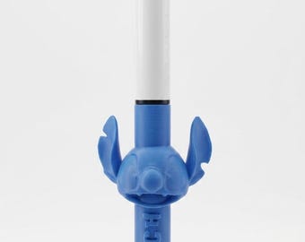 May include: A multi-color ballpoint pen featuring a white upper barrel with a top-mounted selector mechanism and a blue lower barrel. The pen includes a blue molded character topper of Stitch with ears and the word STITCH embossed vertically.