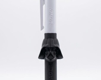 May include: A multi-color Bic ballpoint pen featuring a white barrel with blue and green ink selectors. The lower portion of the pen is encased in a black 3D-printed Darth Vader helmet topper with the text DARK VADER printed vertically.