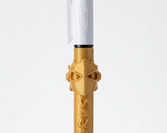 May include: A multi-color retractable ballpoint pen with a white upper barrel and a gold-colored lower barrel. The gold section features a sculpted helmet-like emblem and the embossed text ACTARUS. The top includes blue and green ink selectors.
