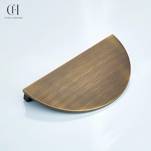 May include: A semi-circular cabinet pull with a brushed antique brass finish and a black base. The hardware features a smooth, curved top surface with a metallic texture. The logo for Citadel Hardware is visible in the upper left corner.