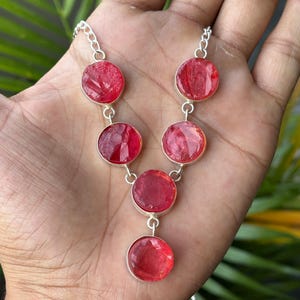 May include: A silver necklace featuring five round, faceted, translucent red stones set in polished metal bezels. The stones are arranged in a symmetrical pattern, with a single central drop stone suspended from the lowest point of the chain.